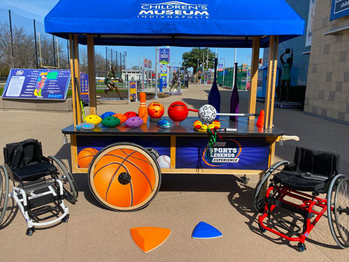 Adaptive sports balls and equipment sitting on a blue cart. A youth sized sport wheelchair is on the left. An adult sized sport wheelchair is on the right.